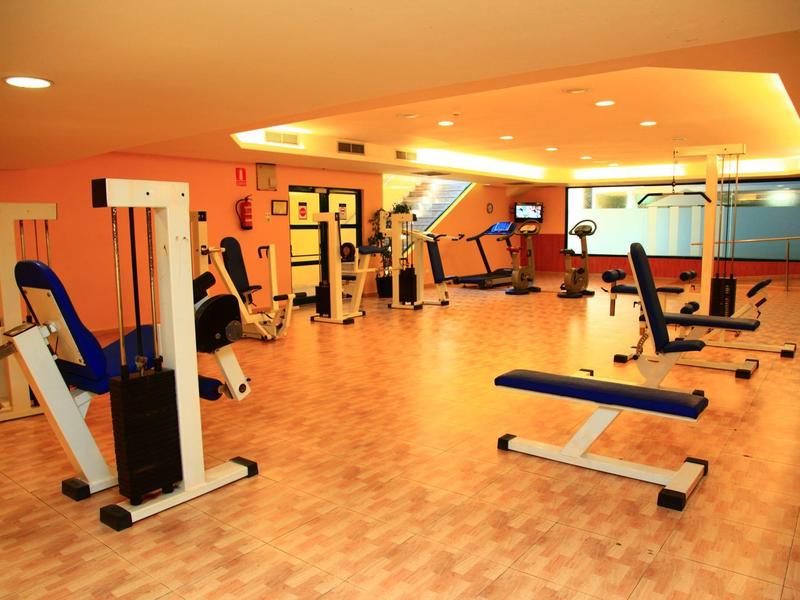 Bright gym with wooden floor, blue workout benches, and white exercise machines.