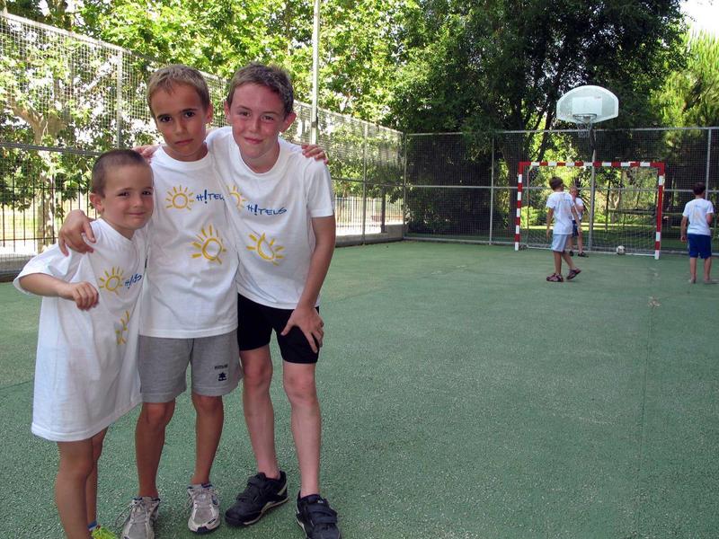 Three children wearing white shirts stand closely together on a green sports court outdoors.