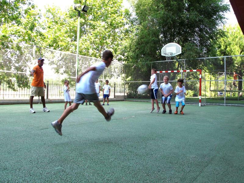People playing soccer on a fenced outdoor sports court surrounded by trees.