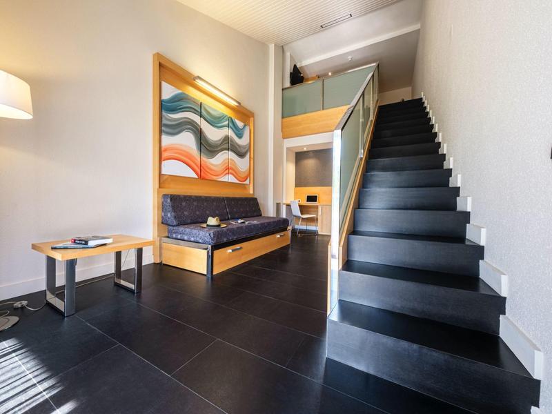Habitación de hotel moderna con escalera negra, cuadro colorido y sofá junto a una mesa redonda de madera.