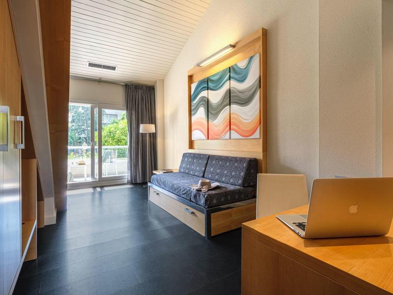 Sala de hotel con sofá cama, ventana grande y escritorio con portátil bajo luz cálida.