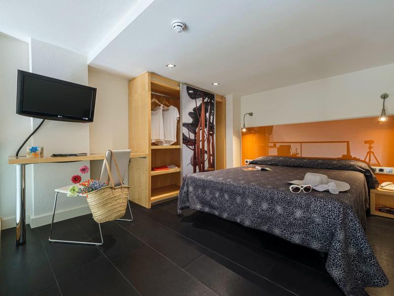 Habitación de hotel moderna con cama doble, televisión montada y armario abierto con ropa colgada.