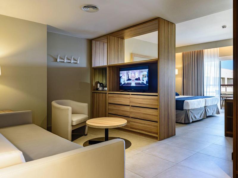 Moderna habitación de hotel con área de estar, armario de madera y vista al dormitorio con cama doble.