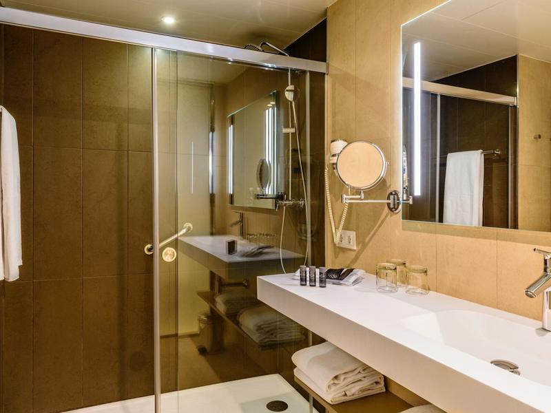 Baño moderno con amplia ducha de vidrio, gran espejo y lavabo largo con grifos.