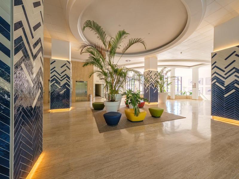 Vestíbulo de hotel luminoso con grandes ventanas, plantas y cojines coloridos en el suelo.