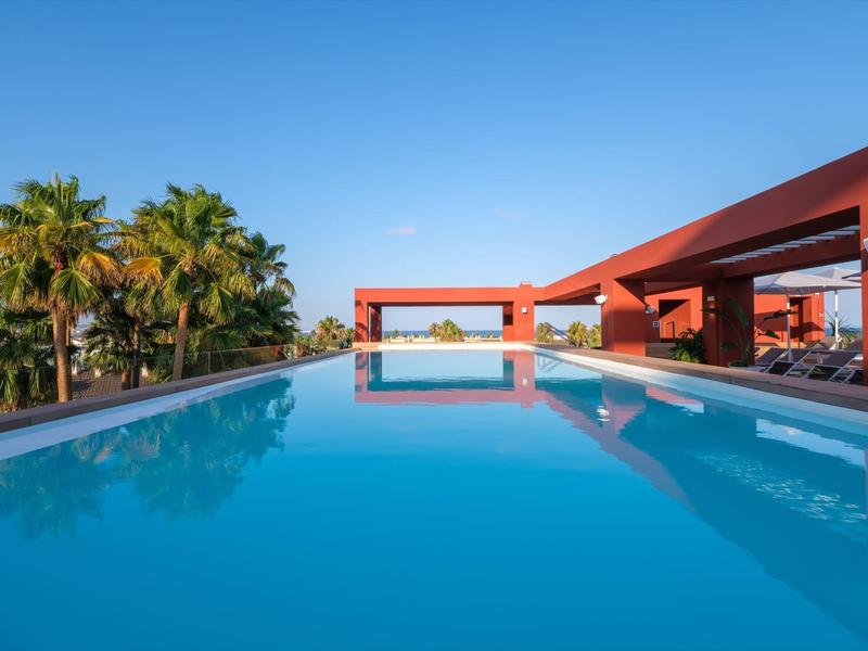 Piscina moderna con agua azul clara junto a una terraza y palmeras bajo un cielo azul.