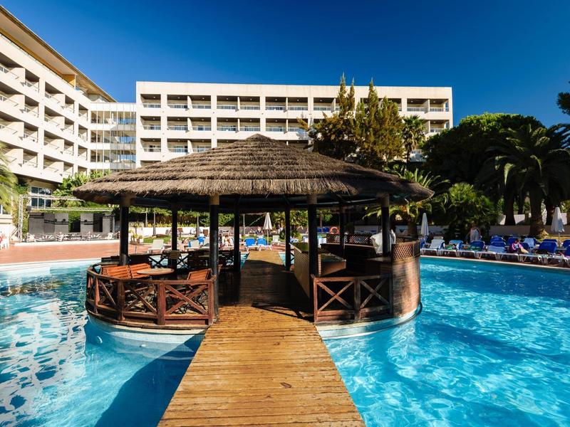 Hotel con piscina al aire libre y bar en isla central con techo de paja bajo cielo despejado.