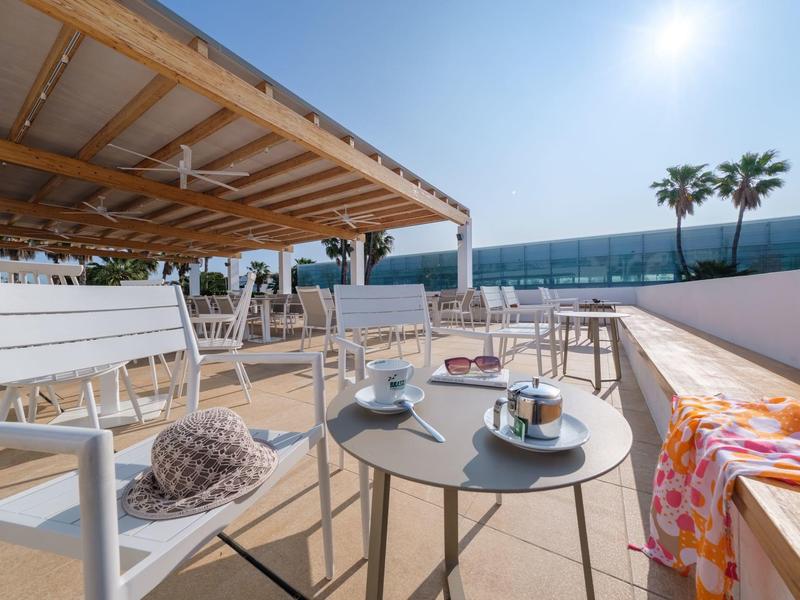 Terraza con mesas y sillas bajo un cielo despejado, vista al mar y palmeras, ambiente de hotel.