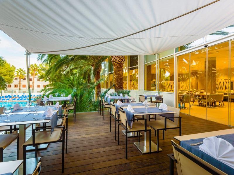 Terraza de restaurante en hotel con mesas y sillas, techo blanco y vegetación alrededor