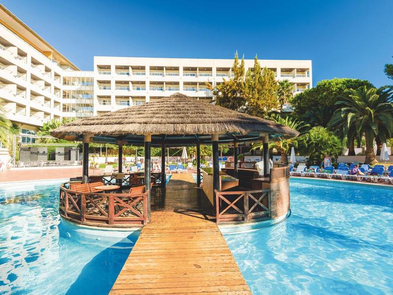 Piscina con bar en isla y camino de madera frente a un hotel grande bajo un cielo azul claro.