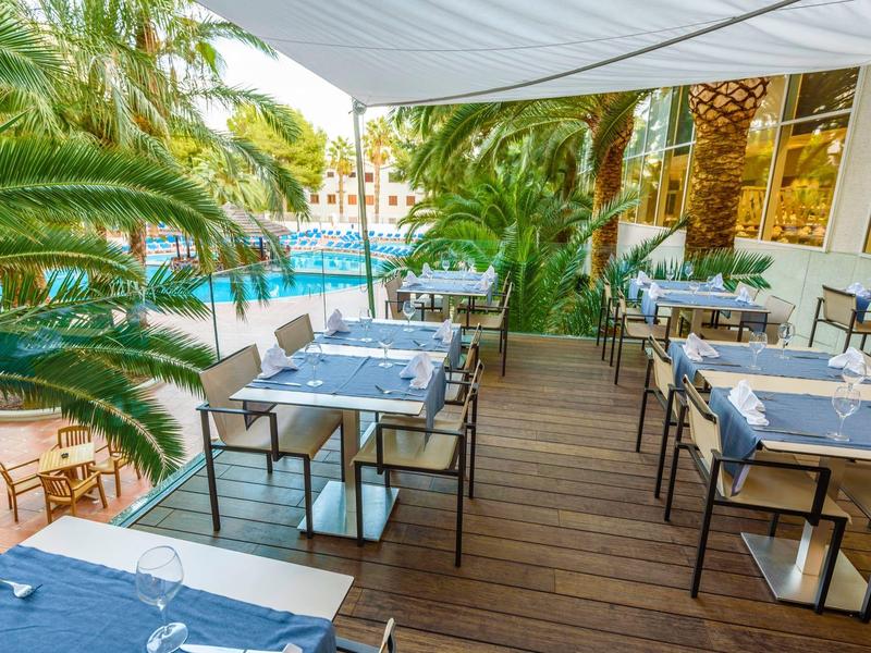 Restaurante al aire libre con suelo de madera y vista a una piscina, rodeado de palmeras y sombrillas.