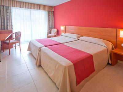 Camera d'hotel con due letti singoli, parete rossa e scrivania vicino alla finestra.