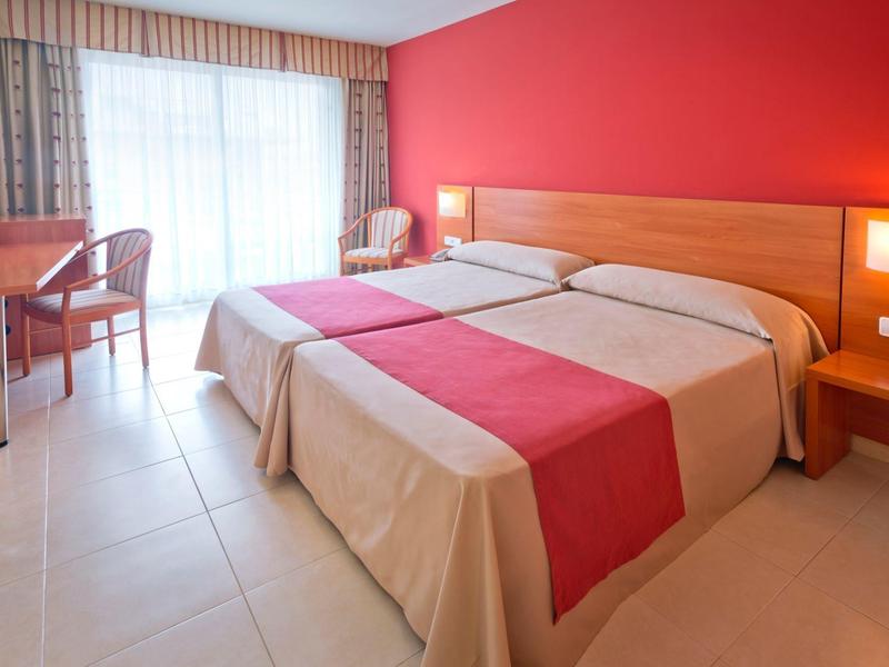 Camera d'hotel con due letti singoli, parete rossa e scrivania vicino alla finestra.