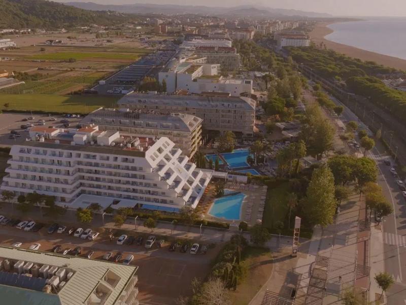 Vista aerea di un hotel costiero con più edifici, una piscina e una strada adiacente.