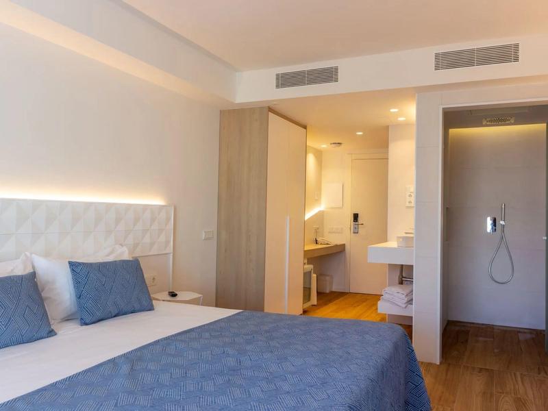 Camera d'hotel moderna con letto matrimoniale, cuscini blu, pavimento in legno e bagno aperto.