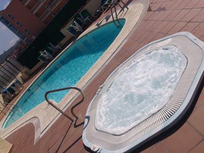 Área de piscina y jacuzzi con agua azul y baldosas de terraza en un jardín de hotel.