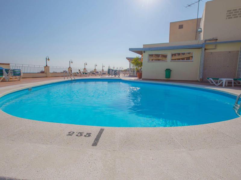 Una piscina ovalada con agua azul clara en una terraza de hotel bajo un cielo soleado.