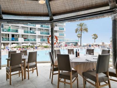 Overdekt zitgedeelte met tafels en stoelen met uitzicht op zwembad en hotelgebouw.