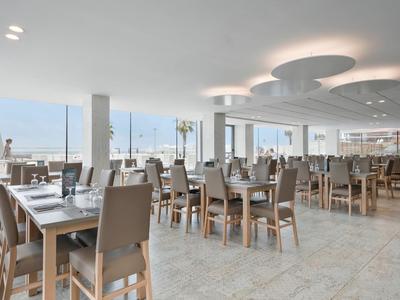 Licht, modern restaurant met houten stoelen en tafels, grote ramen met uitzicht op zee.