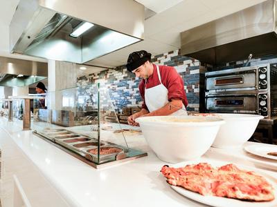 Kok bereidt eten in open restaurantkeuken met pizza en grote witte kommen.