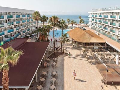 Uitzicht op hotel met zwembad en zee op de achtergrond, omgeven door palmbomen en parasols.