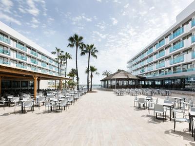 Ruimtelijk buitengedeelte van hotel met tafels, stoelen en palmbomen onder bewolkte lucht.