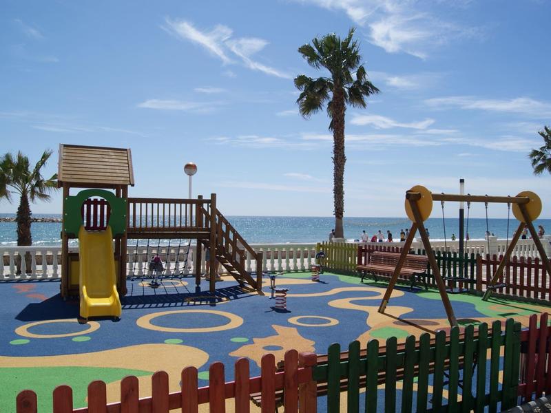 Kleurrijke speeltuin bij het strand met glijbaan, schommel en palmbomen op de achtergrond.