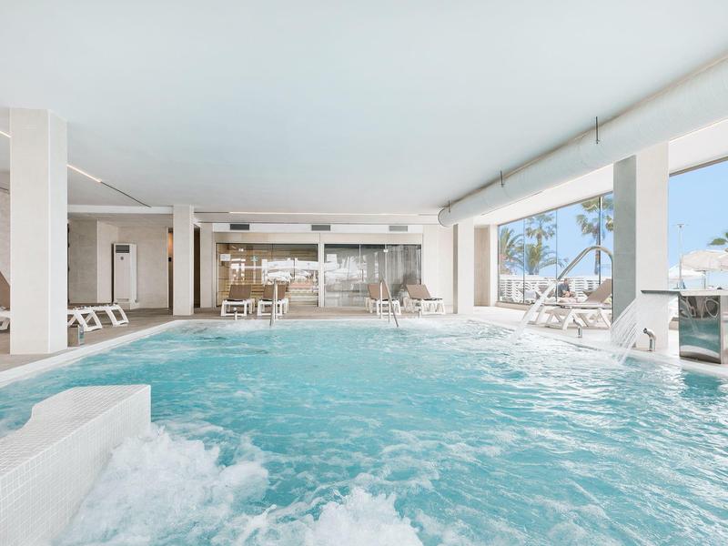 Licht binnenzwembad met jacuzzi, ligstoelen en grote ramen met uitzicht op zee.