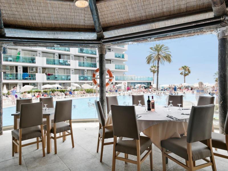 Overdekt zitgedeelte met tafels en stoelen met uitzicht op zwembad en hotelgebouw.