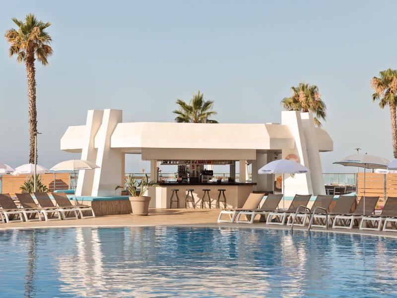 Moderne zwembadomgeving met ligstoelen, bar en palmbomen onder een heldere lucht.