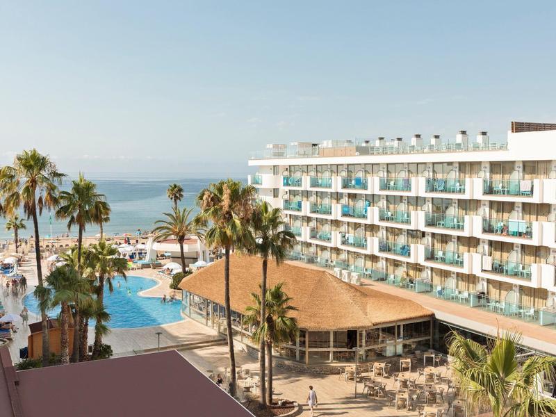 Hotel met zwembad, palmbomen en zeezicht op een zonnige dag.