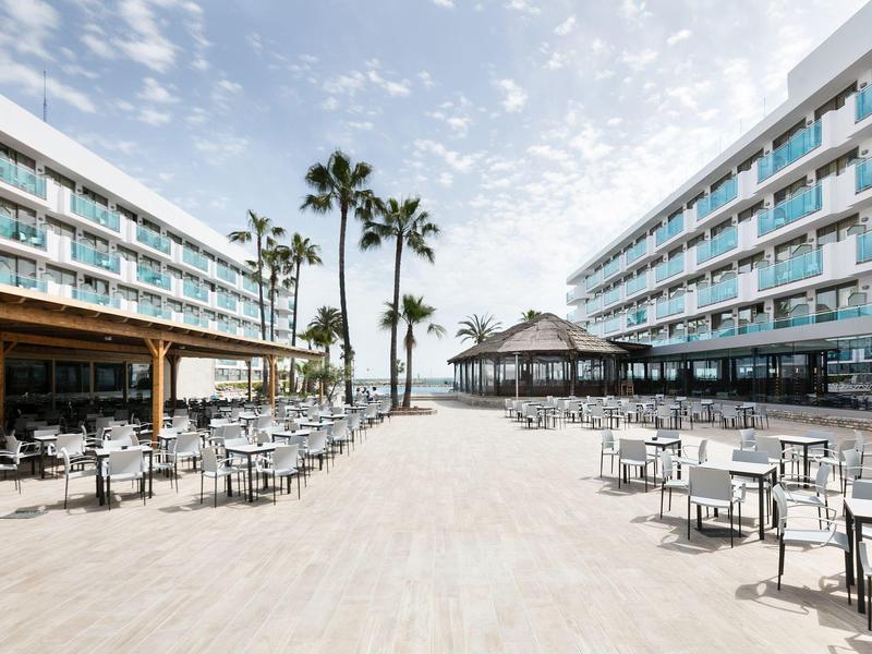 Ruimtelijk buitengedeelte van hotel met tafels, stoelen en palmbomen onder bewolkte lucht.