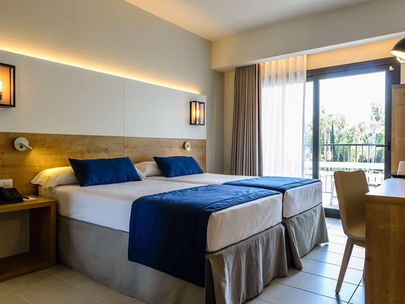 Modern hotel room with double bed, blue accents, and balcony overlooking greenery.