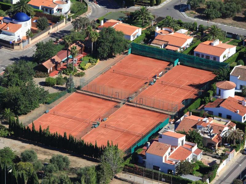 Four adjacent red tennis courts in a residential area with white houses.