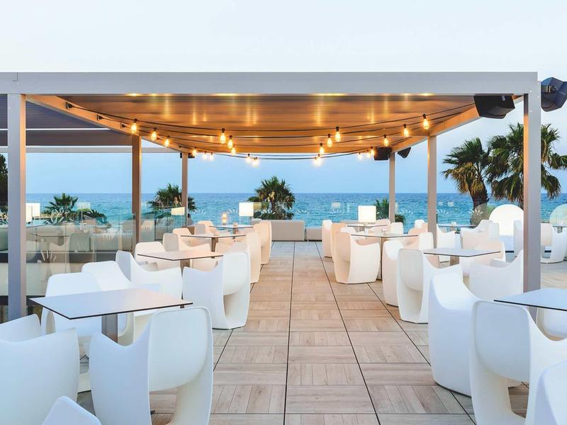 Open dining area with white chairs and tables under a wooden roof with string lights by the sea.