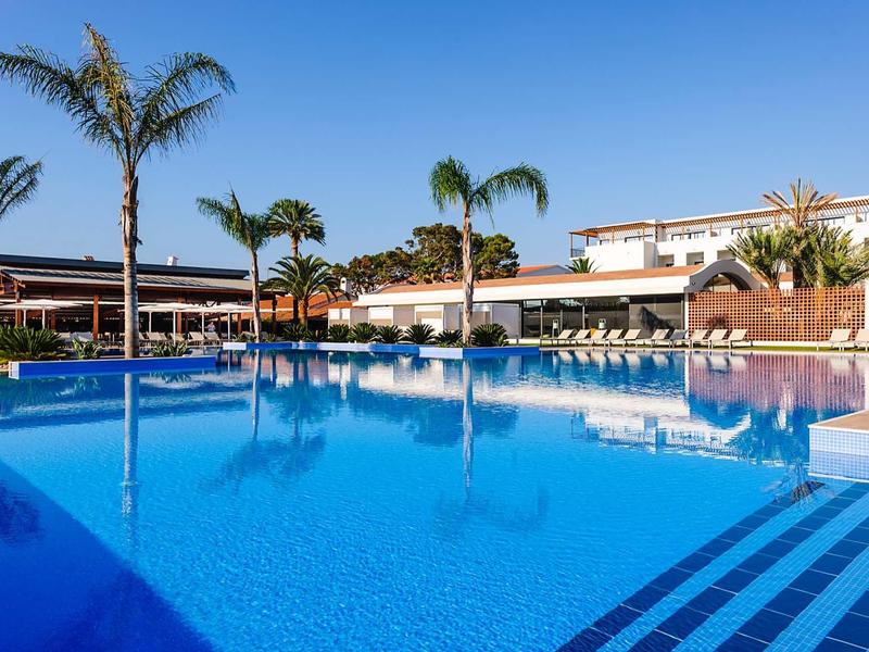 Large outdoor pool at a hotel with palm trees and sun umbrellas around the edge.