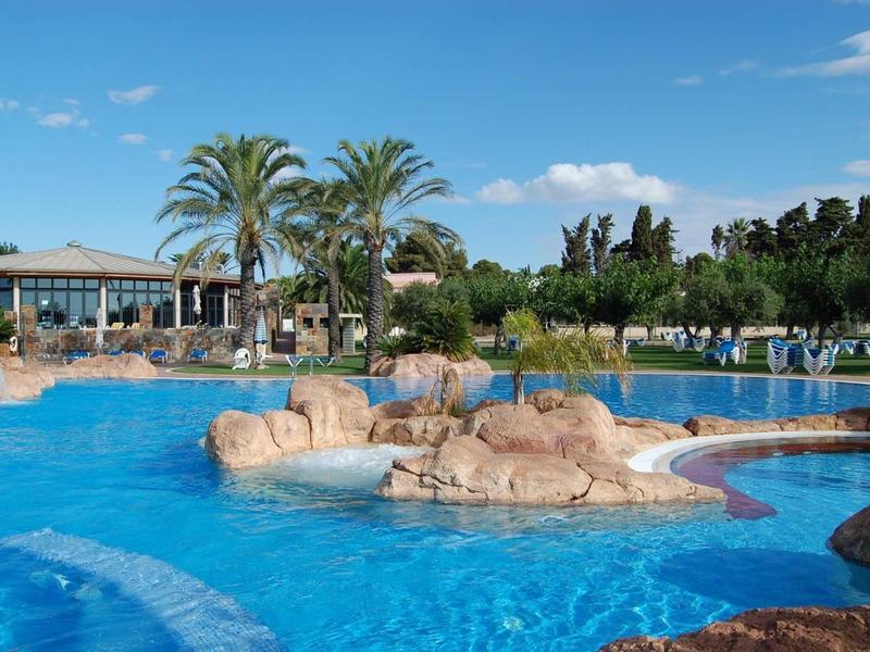 Large hotel pool with rocks and palm trees under a blue sky with few clouds.