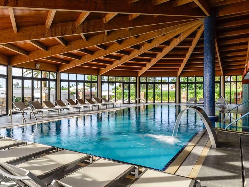 Indoor pool with wooden beam ceiling and lounge chairs by large windows overlooking greenery.
