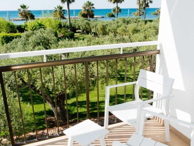 Balcony with white chairs and table, view of palm trees, green garden, and sea in the background.