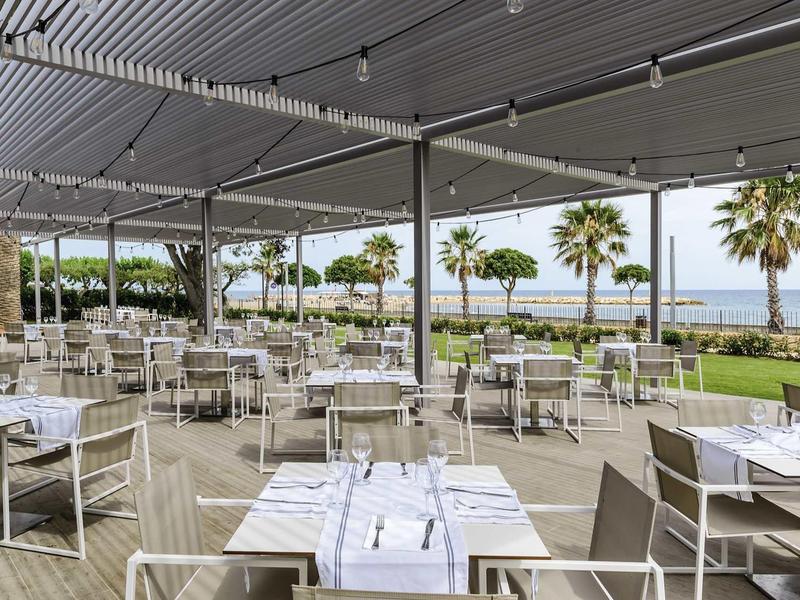 Open dining area with white tables and chairs under a large canopy overlooking palm trees and the sea.