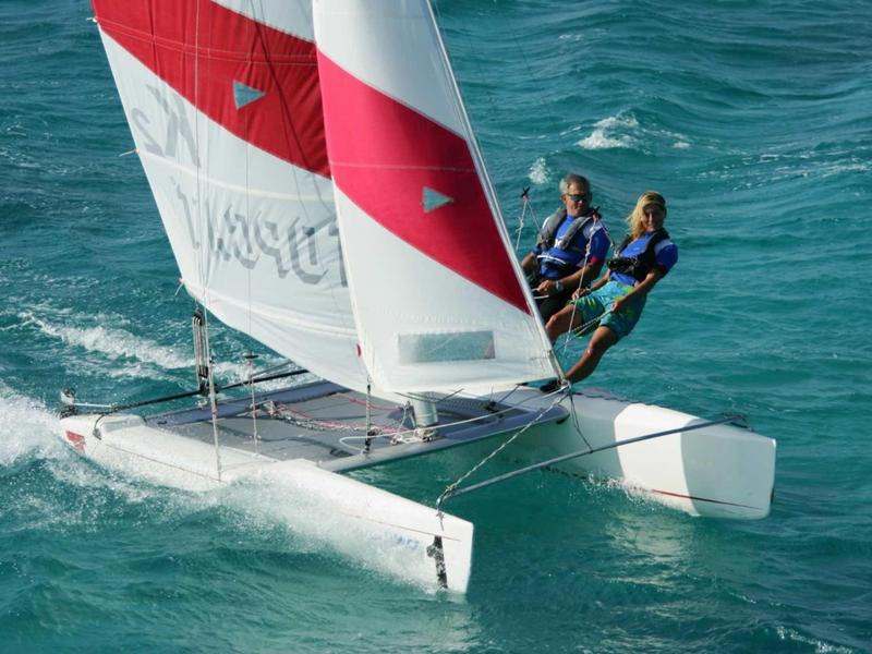 Two people sailing a catamaran with red and white sails on blue water.