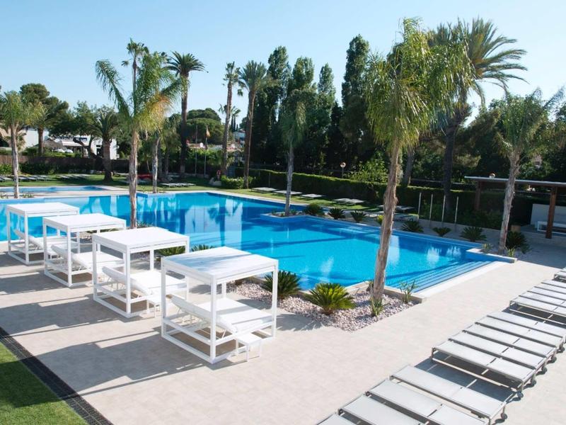 Modern hotel pool with white lounge chairs and tables surrounded by palm trees.