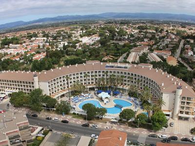 Gran hotel curvado con piscina, rodeado de árboles y paisaje urbano al fondo.