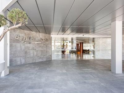 Entrada moderna de un hotel con baldosas grises y paredes de vidrio bajo un techo metálico.