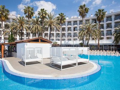 Hotel mit großem Pool, weißen Tagesbetten und Palmen vor einem mehrstöckigen Gebäude