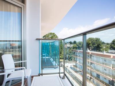 Balcón con silla blanca, barandilla de vidrio y vista a edificios y árboles bajo cielo azul.