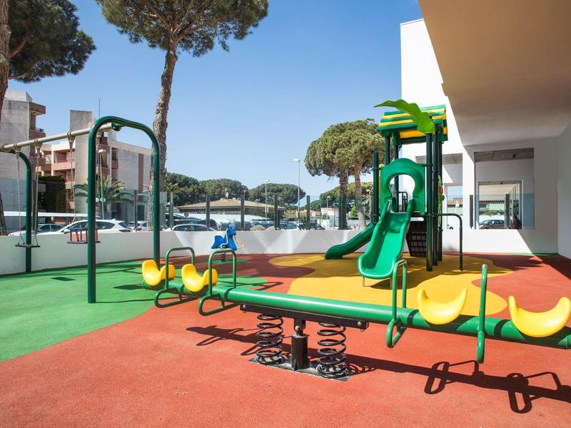 Área de juegos para niños colorida con toboganes y columpios junto a un edificio de hotel bajo el cielo azul.