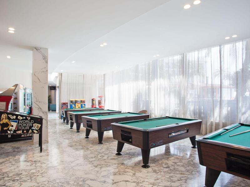 Interior con varias mesas de billar y suelo de mármol claro, cortinas claras en las ventanas.