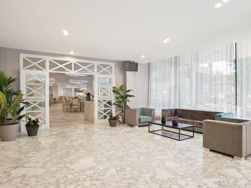 Área de recepción luminosa con asientos, grandes ventanas y plantas en macetas.