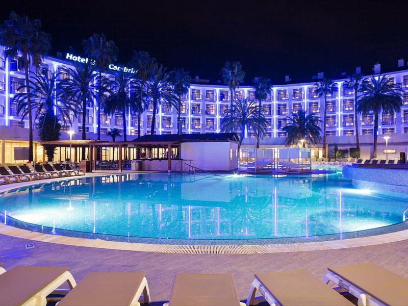 Piscina iluminada del hotel con tumbonas y palmeras frente a un edificio de varios pisos por la noche.